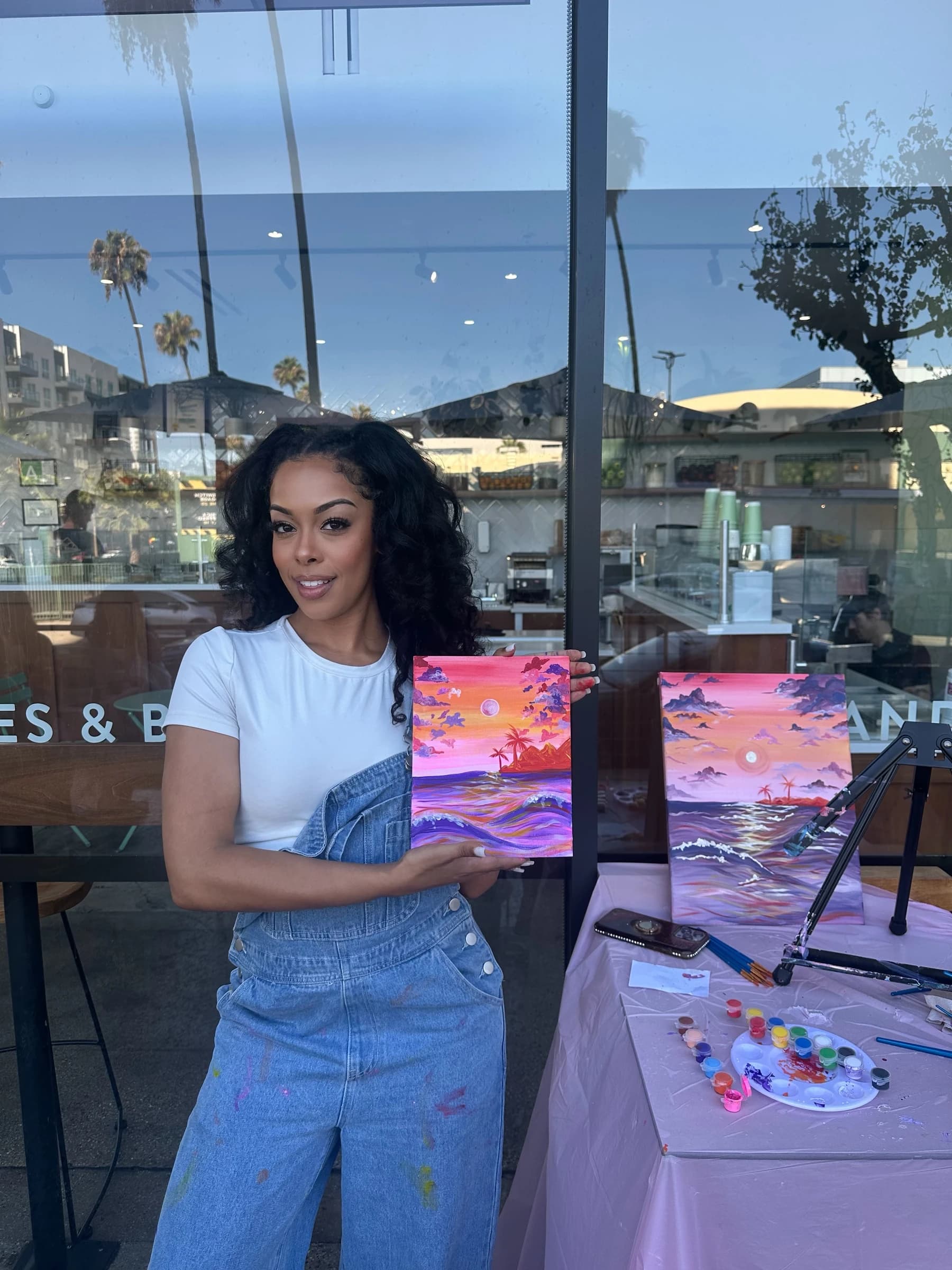 Aliyah Celeste holding a colorful sunset painting beside another canvas and painting supplies at an outdoor art event.