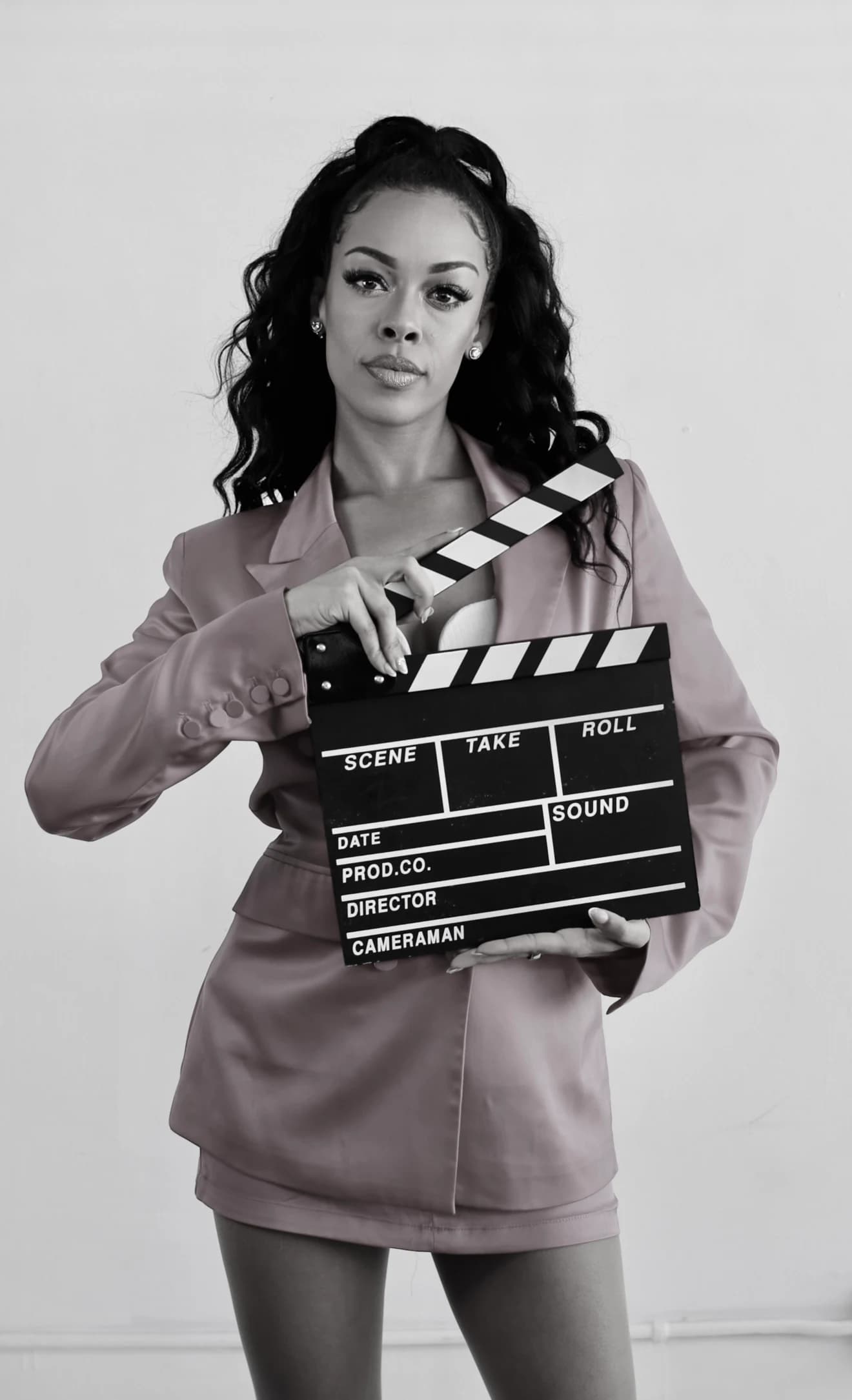 Aliyah Celeste holding a clapperboard, wearing a muted mauve suit.